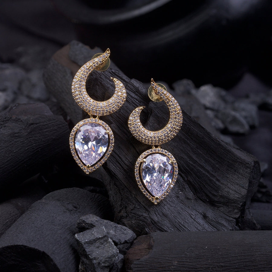 Gold-plated earrings with clear teardrop gemstones resting on black charcoal pieces