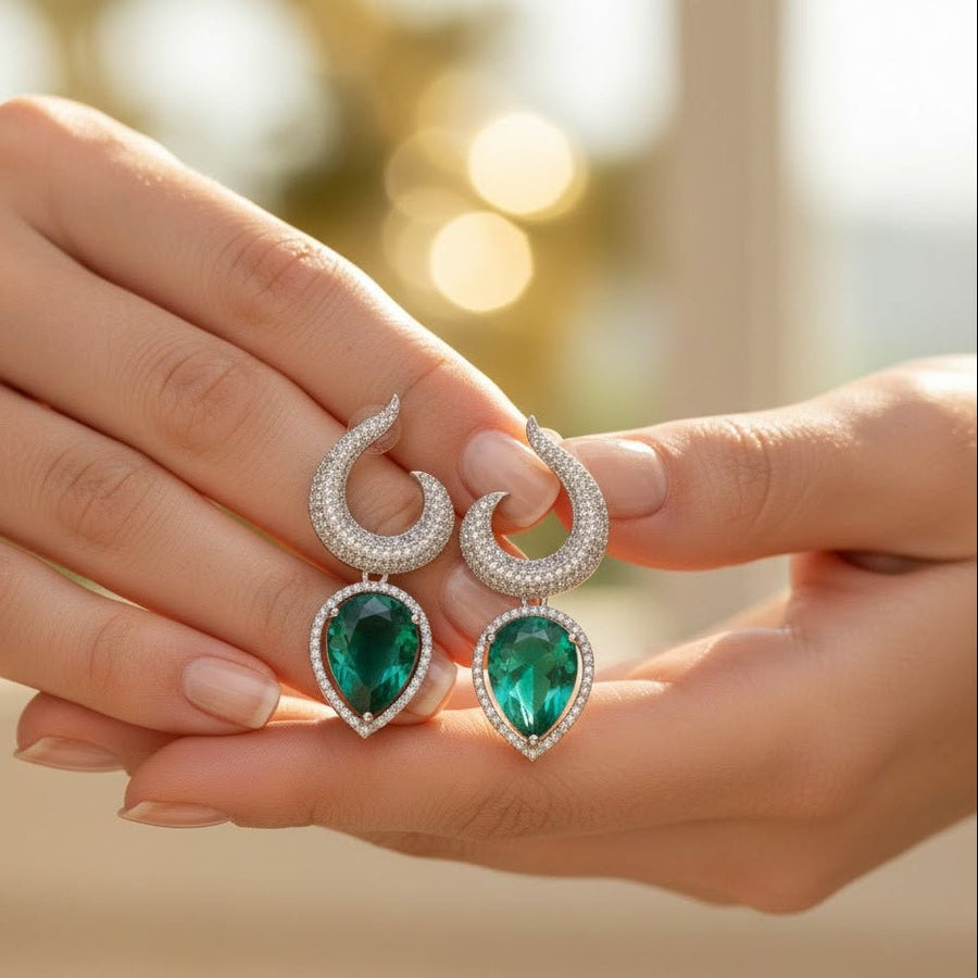 Close-up of woman holding a pair of elegant silver-plated earrings with green teardrop gemstones and crystal-studded swirl tops