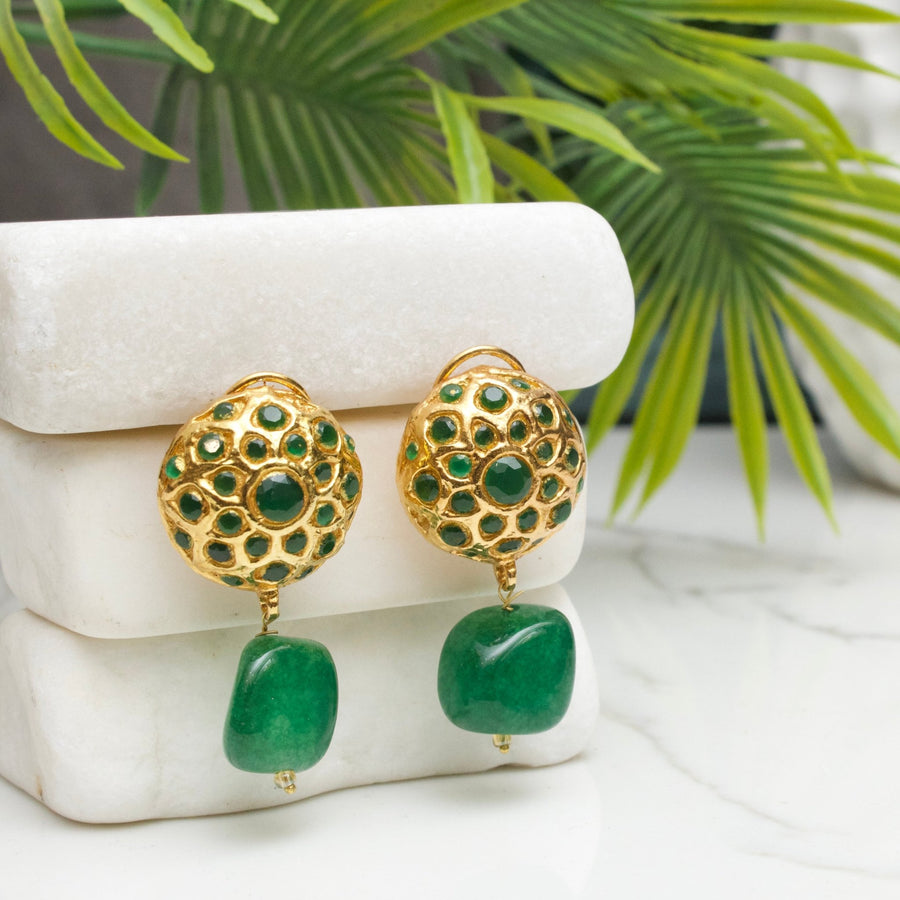 Pair of gold-tone drop earrings with intricate cut-out design and green gemstone drop, displayed on white stone blocks with green foliage background.