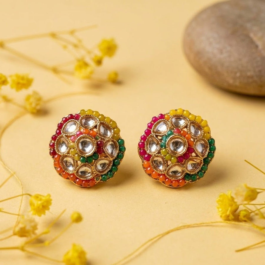 Close-up of gold-plated round stud earrings featuring a floral-inspired pattern with multi-color beads and clear Polki-style stones on a beige surface with dried flowers