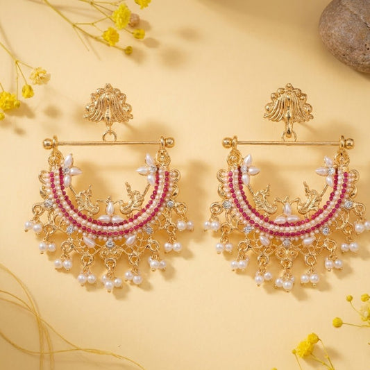 Pair of gold-plated jhumka earrings with ruby red stones and pearl hangings displayed on cream background