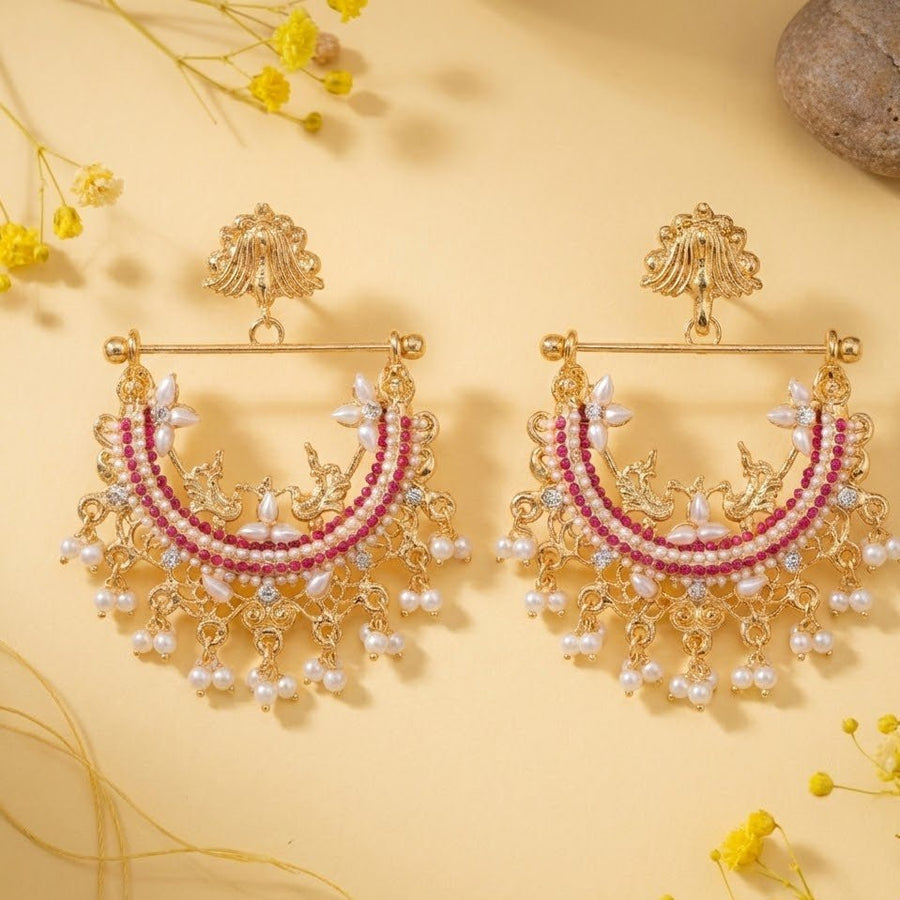 Pair of gold-plated jhumka earrings with ruby red stones and pearl hangings displayed on cream background