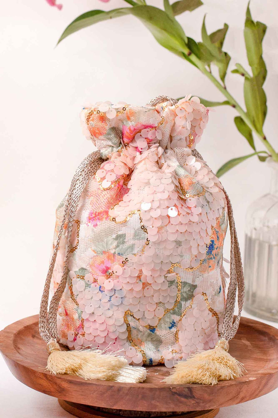Pastel floral embroidered potli bag adorned with pink sequins and rose gold tassels, placed on a wooden plate with a decorative plant background.