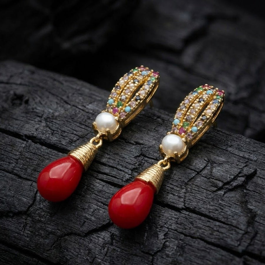 Pair of gold-tone earrings with colorful crystal detailing, central pearls, and vivid red teardrop drops on a dark textured surface