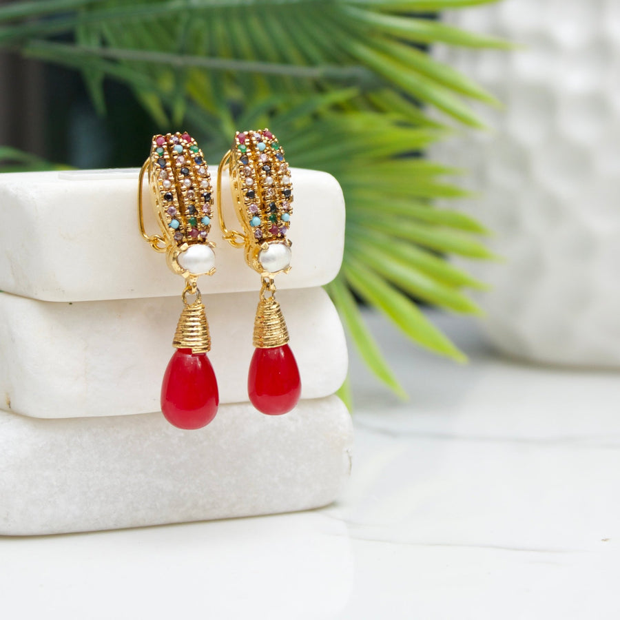 Elegant red teardrop pearl earrings with multicolor crystals placed on stacked white stone slabs with greenery blurred in background