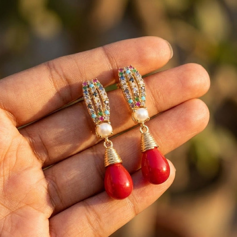 Hand holding a pair of gold-tone earrings with pearl accents and bright red teardrop pendant highlighting colorful crystal details