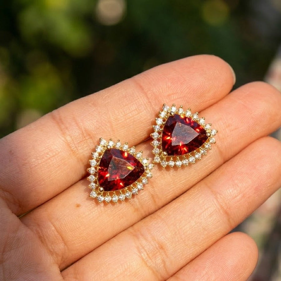 Hand holding deep red trillion cut stud earrings with clear halo stones, showcasing size and brilliance outdoors