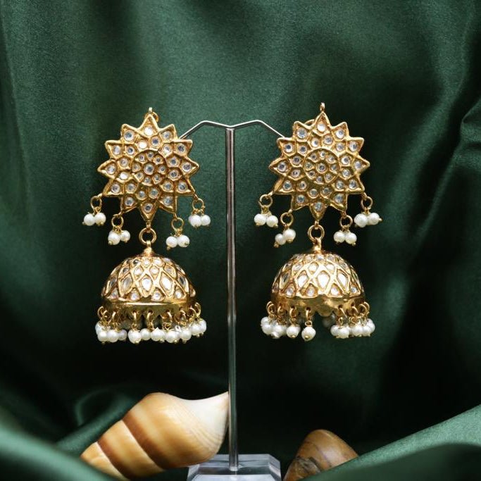 Pair of gold-plated jhumka earrings with floral stone designs and hanging pearl beads displayed on a clear stand against green fabric