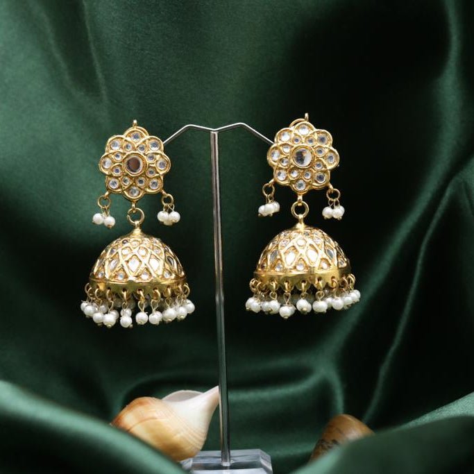 Pair of gold-plated Jhumka earrings with flower-shaped studs, embedded stones, and dangling pearl clusters displayed on stand