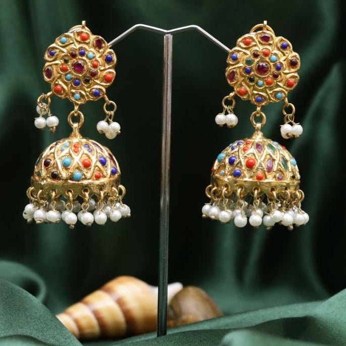 Pair of gold plated Jhumka earrings adorned with multicolored stones and pearl dangles displayed on a stand against a green fabric background
