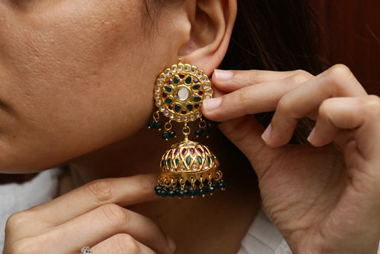 Close-up of a woman wearing gold-plated Jhumka earrings with intricate stonework and black bead tassels, adjusting the earring gently