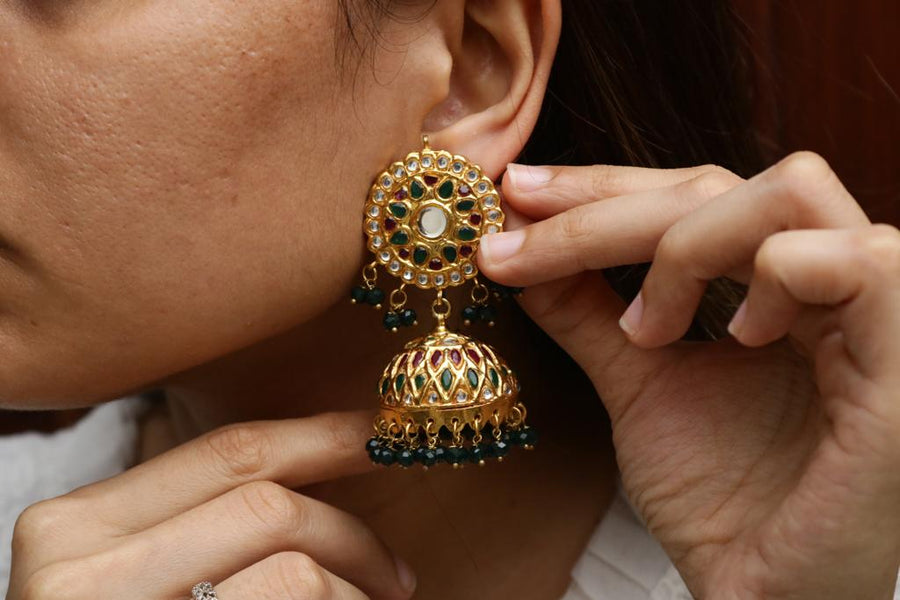 Close-up of a woman wearing gold-plated Jhumka earrings with intricate stonework and black bead tassels, adjusting the earring gently