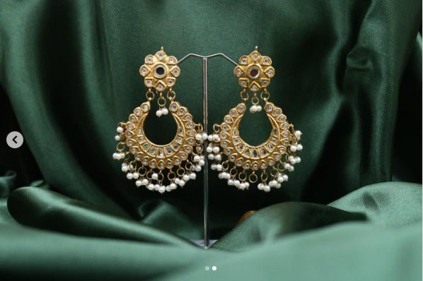 Pair of elegant gold-plated Jhumka earrings adorned with pearls and clear stones displayed on a green satin fabric backdrop