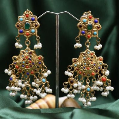 Pair of gold-plated chandelier earrings adorned with colorful stones and dangling pearl clusters displayed on a stand against a green fabric background