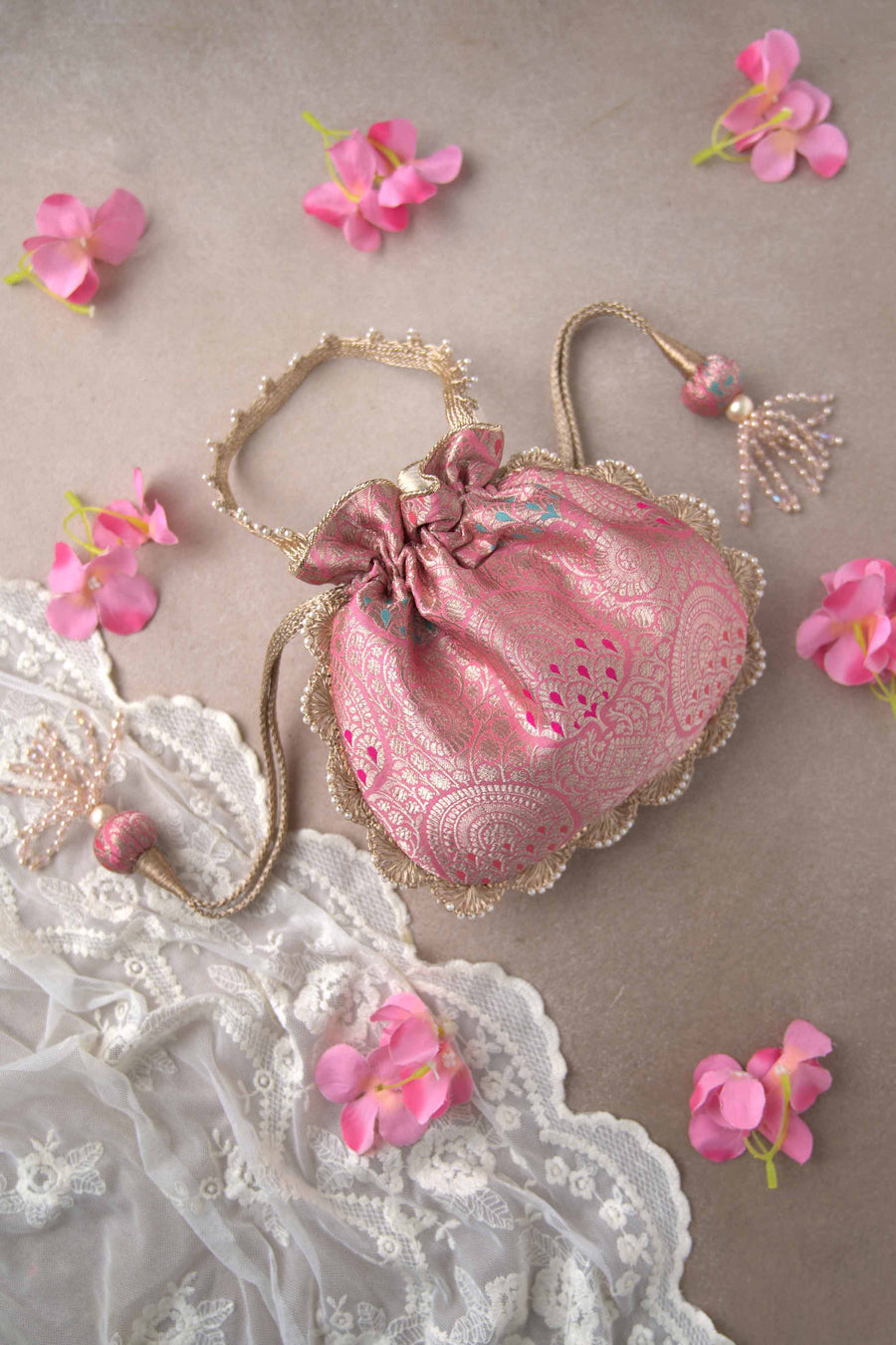Soft pink Banarasi silk potli bag with golden zari floral patterns and beaded tassels, placed on a lace fabric background with pink flowers.