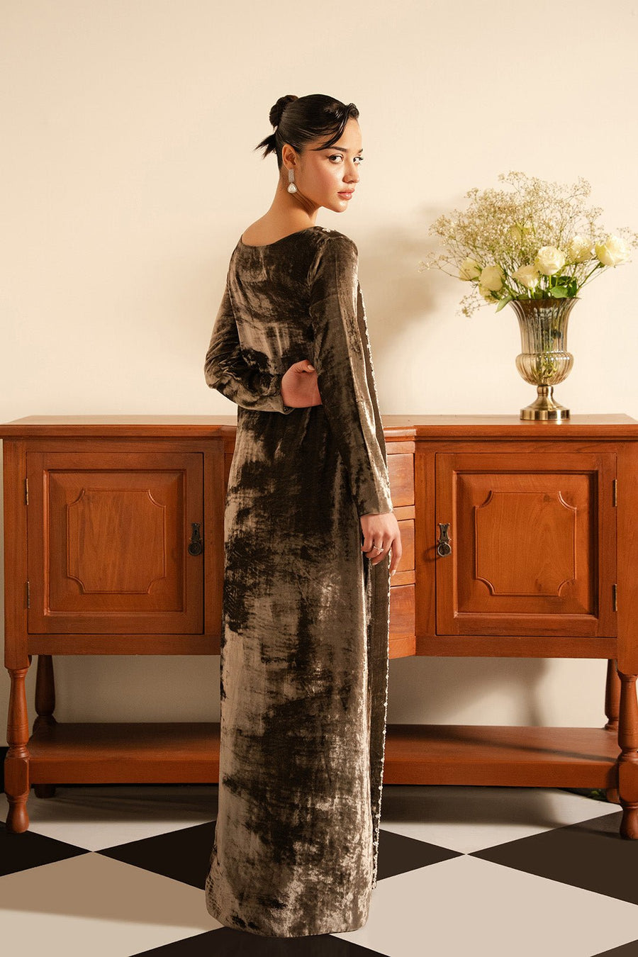 Rear angled view of a woman wearing a floor-length mud grey velvet dress standing in a room with classic wooden furniture and a vase with flowers