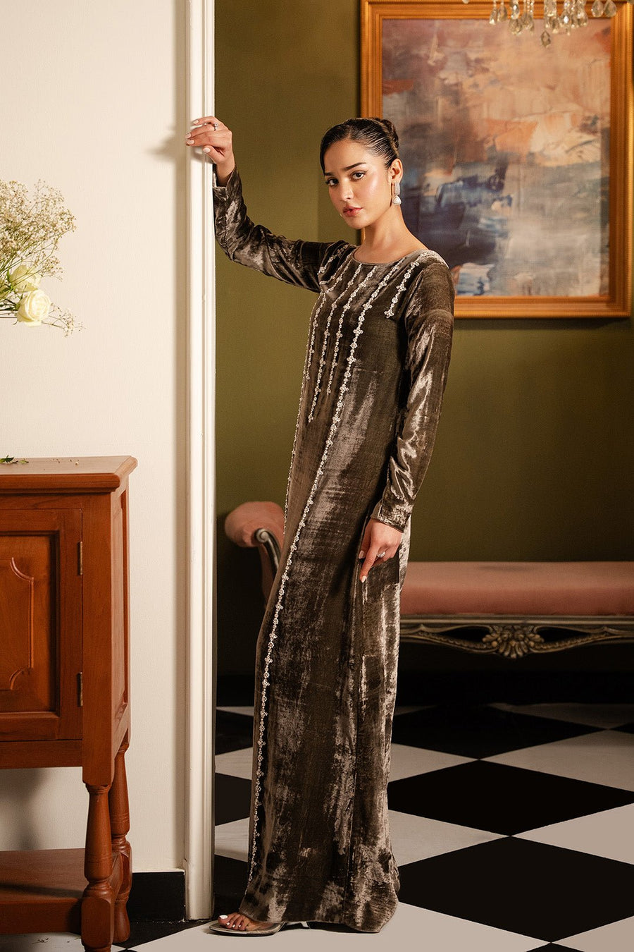 Full side view of a woman in a mud grey velvet dress with vertical embellishments, standing next to a wooden furniture piece in a warmly lit room
