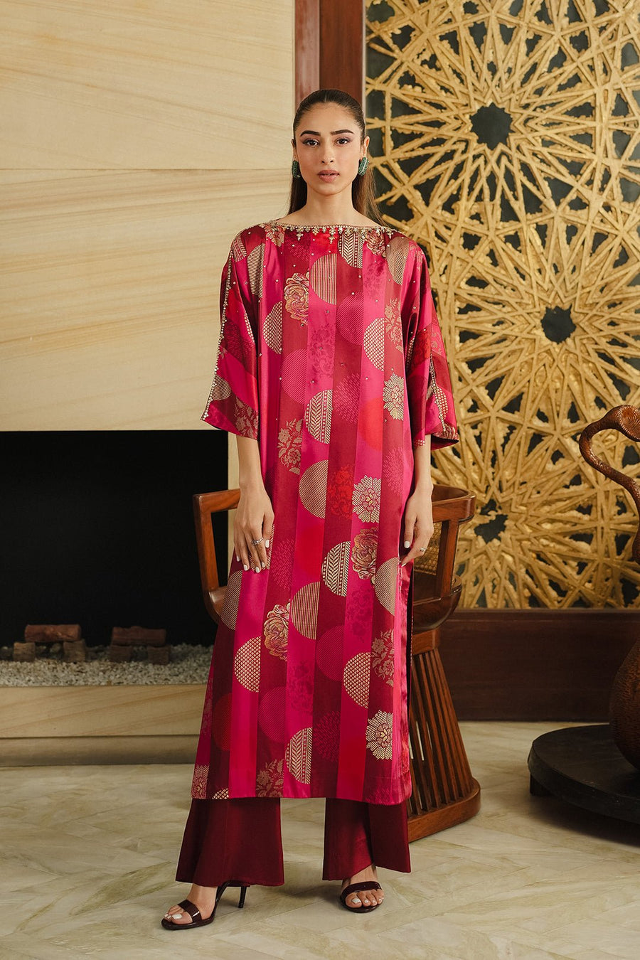 Front view of a woman wearing a long loose-fitting red and burgundy silk dress with gold circular and floral patterns, paired with burgundy wide-leg pants and black heeled sandals, standing indoors.