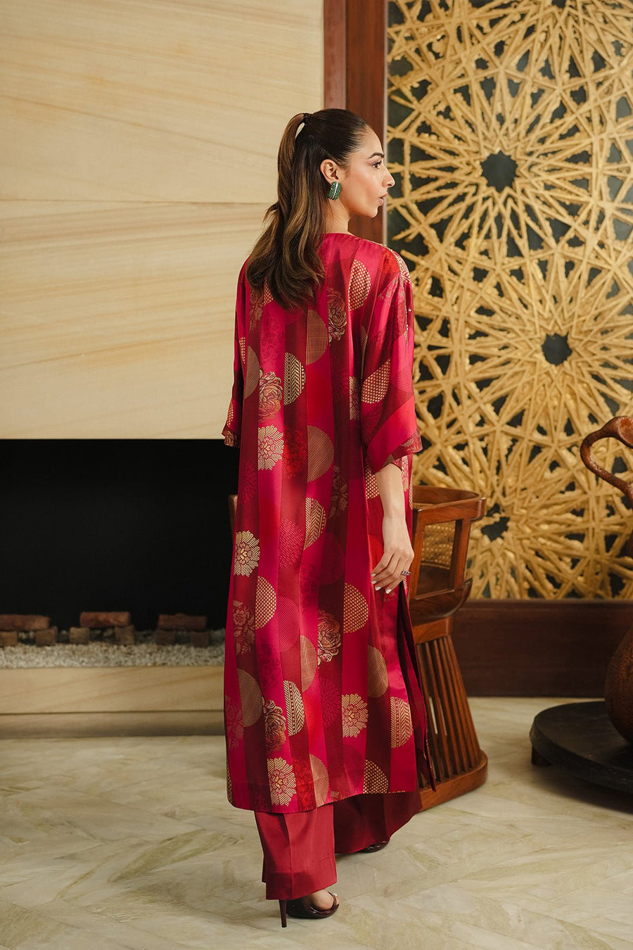 Rear view of the long silk dress with gold patterns showing the loose fit and full-length coverage, worn by a woman with a ponytail standing indoors near wooden furniture.