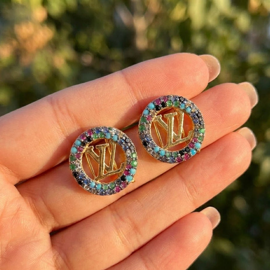 Hand holding a pair of round gold-tone earrings with multicolor stones and central logo outdoors