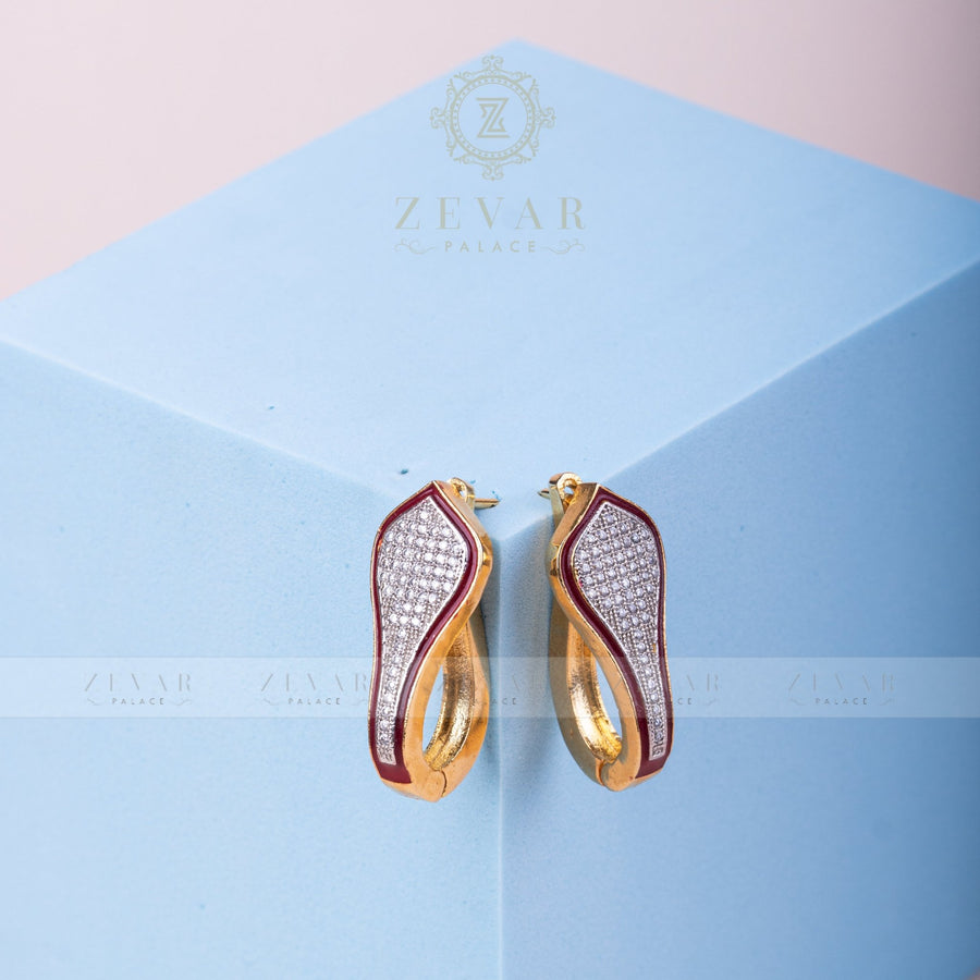 Pair of gold tone teardrop-shaped hoop earrings with maroon enamel border and pavé stone detailing displayed on light blue geometric stand, alternate angle