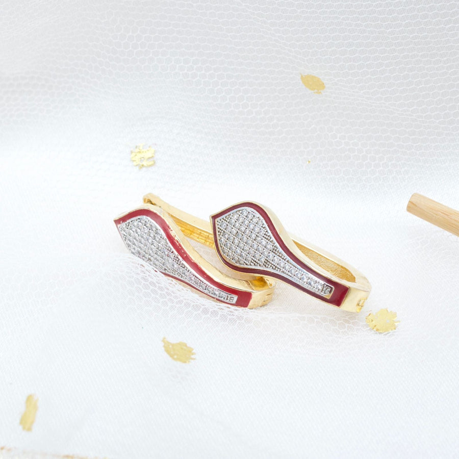 Pair of gold tone teardrop-shaped hoop earrings with red enamel border and pavé stone detailing displayed on light blue geometric stand