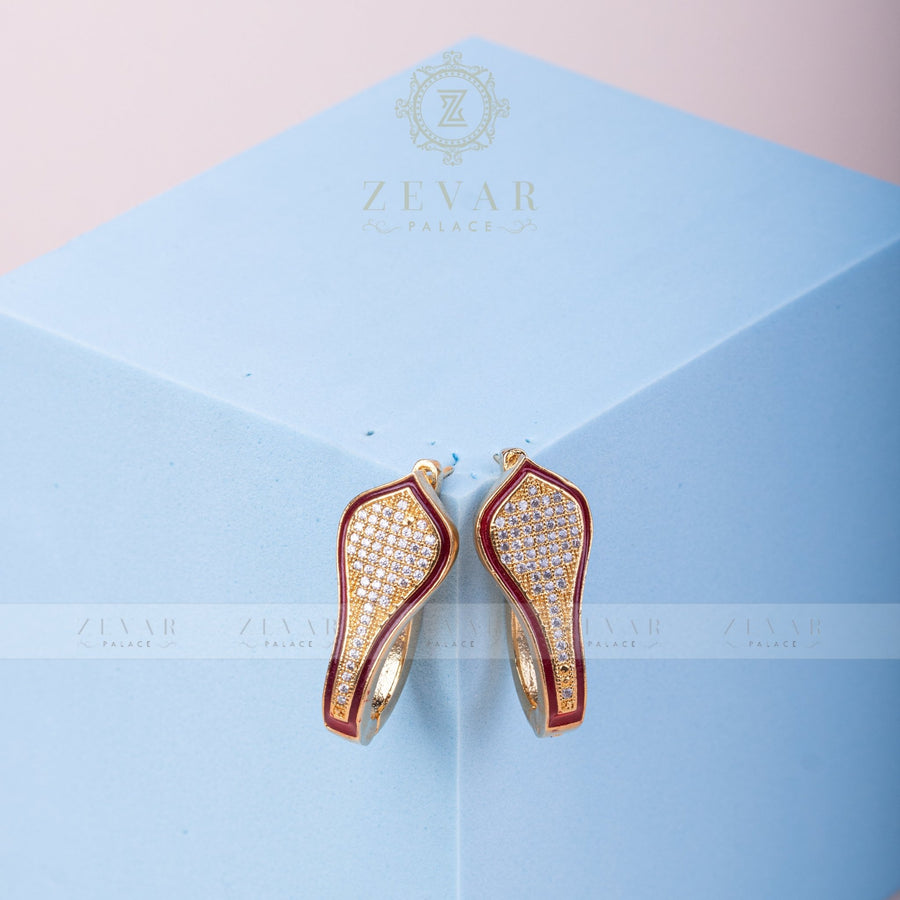 Pair of gold tone teardrop-shaped hoop earrings with maroon enamel border and pavé stone detailing displayed on light blue geometric stand