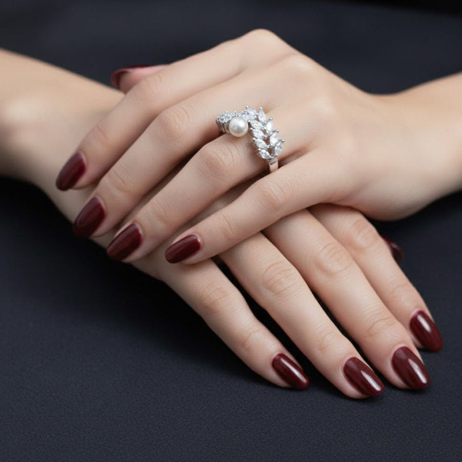 Woman's hand wearing a silver ring with an Akoya pearl and zirconia leaf cluster, set against a black background with dark red nail polish.