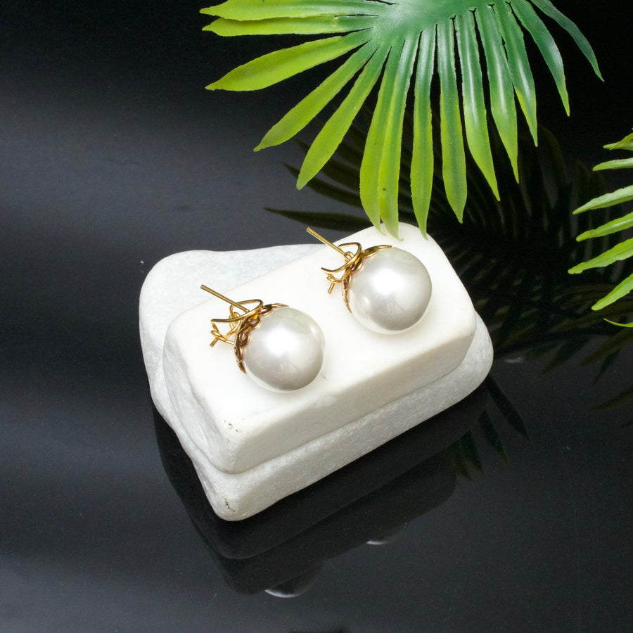 Pair of white pearl stud earrings with gold-tone posts displayed on a white soapstone stand, set against a black glossy background with green palm leaves.
