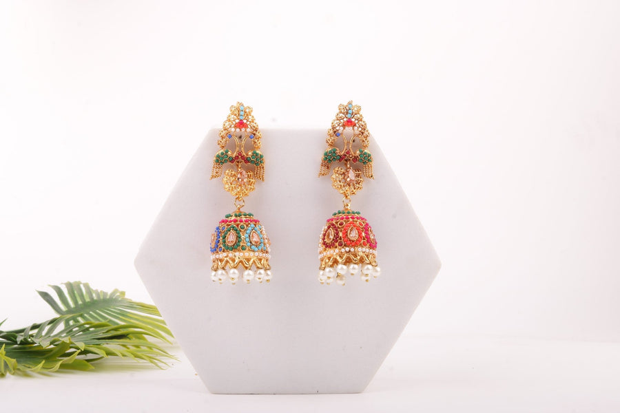Front view of colorful jhumka earrings with detailed beadwork and pearl accents on a white hexagonal display with green leaves nearby