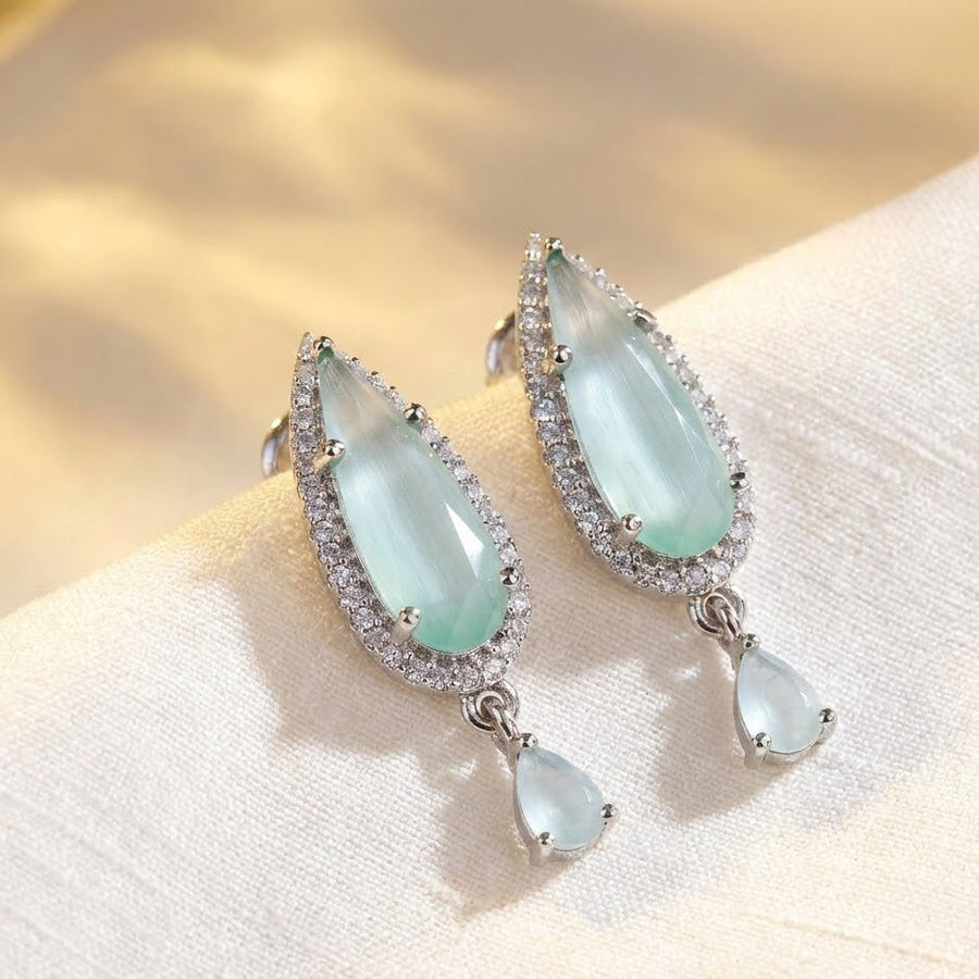 Light aqua teardrop crystal earrings with clear stone halo resting on soft cream fabric background