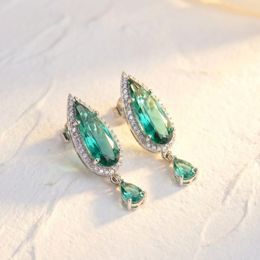 Close-up of green teardrop crystal earrings surrounded by clear stones with small dangling crystal on light textured surface