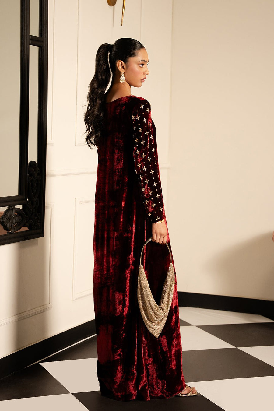 Rear view of woman in maroon velvet dress holding a gold-toned handbag, showing intricate sleeve embellishments and rich texture.