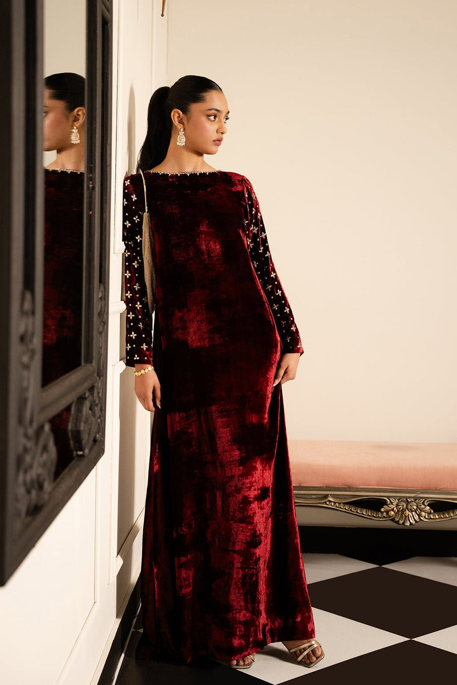 Woman wearing a deep maroon velvet floor-length dress with embellished sleeves standing sideways against a wall.