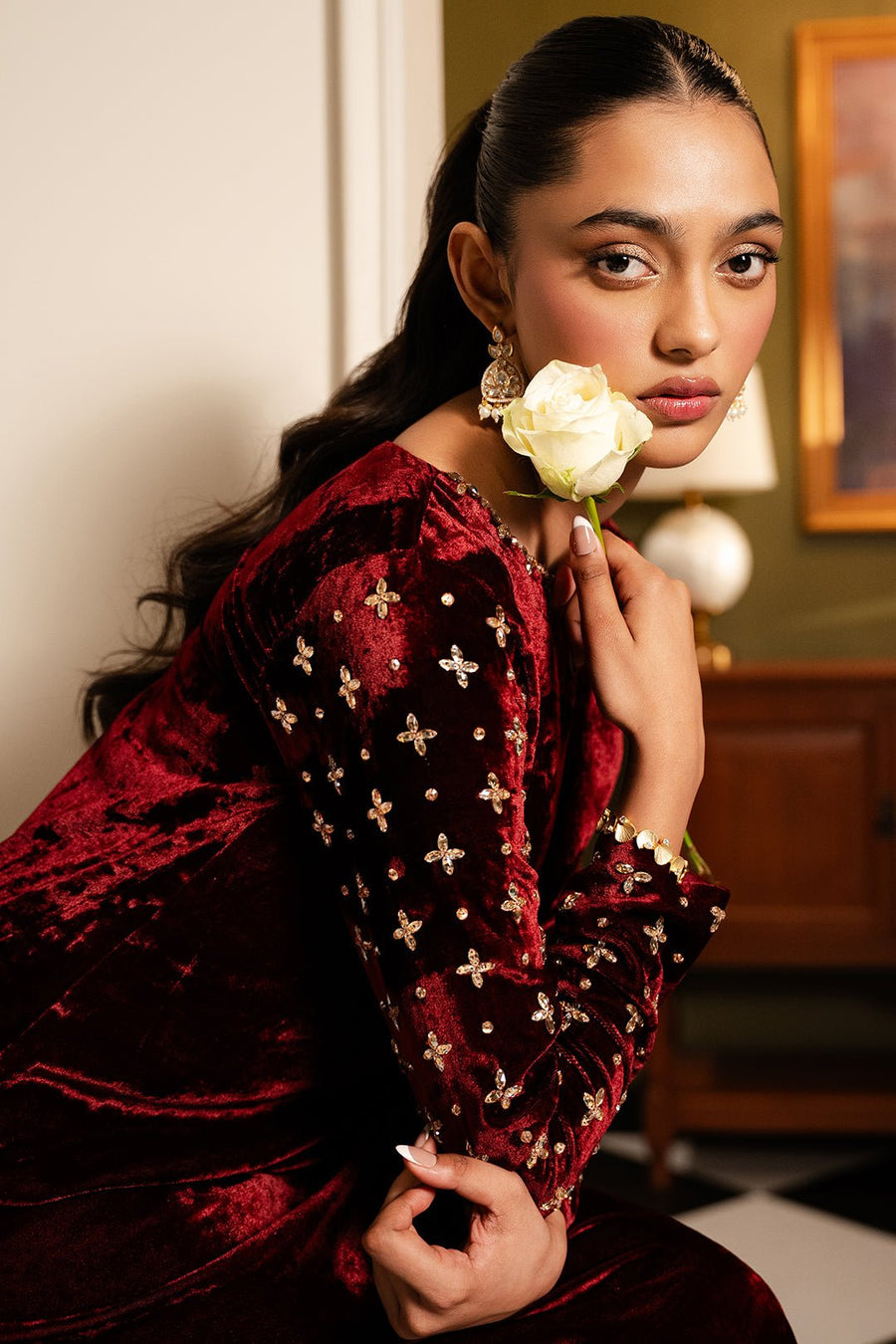 Close-up of a woman in a maroon velvet dress with gold-jewel floral embellishments on the sleeves holding a white rose.