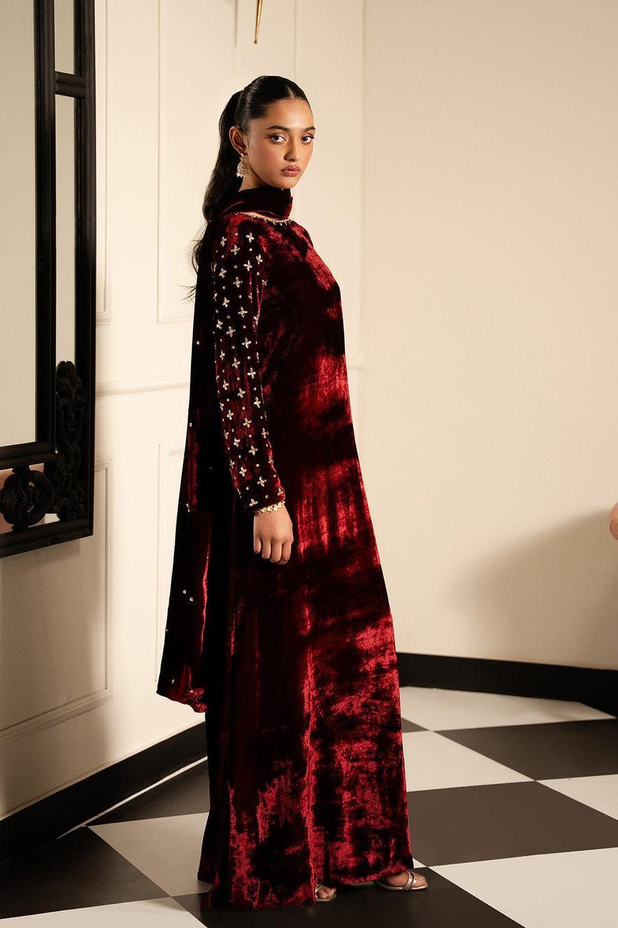 Side view of woman wearing maroon velvet dress with embellished sleeves and high neckline posing in an elegant room.