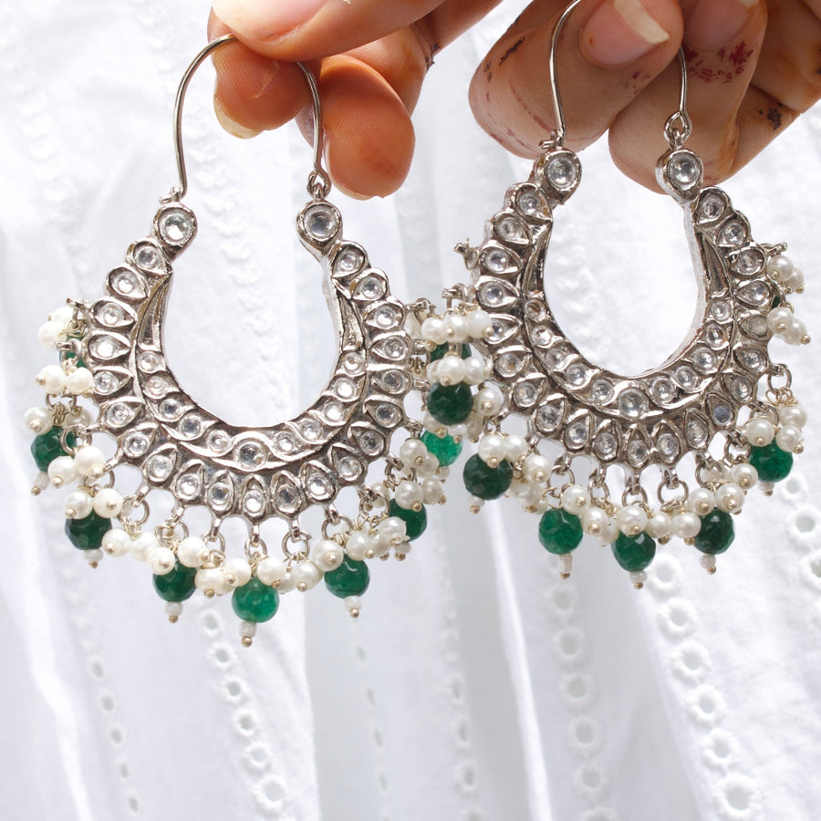 Close-up of traditional silver chandelier earrings held between fingers, featuring clear stones, small white pearls, and green beads.
