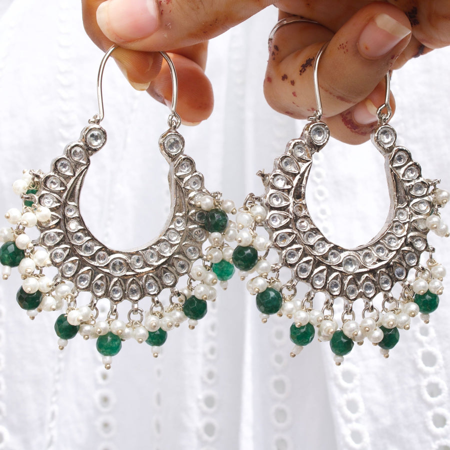 Detailed front view of silver chandelier earrings with pearl and green bead clusters against a white embroidered fabric background.