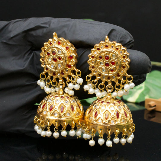 Gold-plated Jhumka earrings with red and clear stones and pearl drops held in a gloved hand
