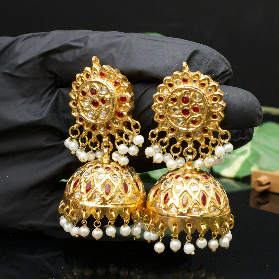 Gold-plated Jhumka earrings with red and clear stones and pearl drops held in a gloved hand