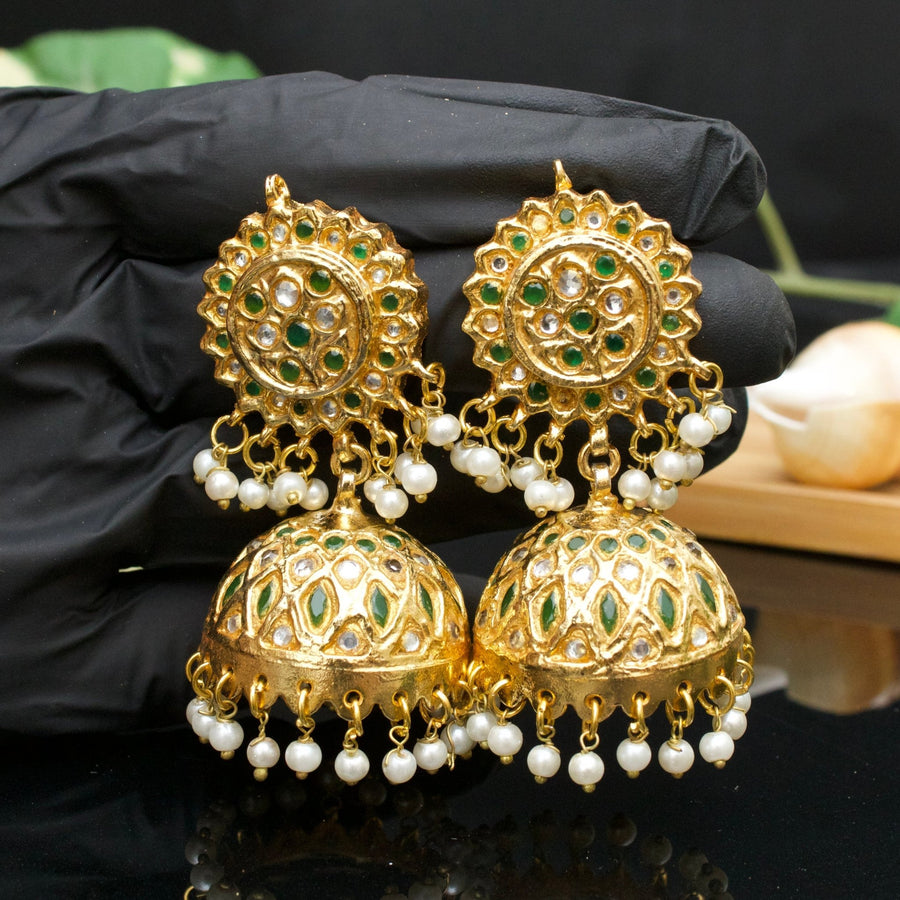 Gold-plated Jhumka earrings with green and clear stones and pearl drops held in a gloved hand