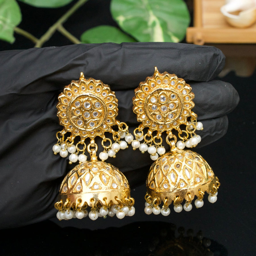 Close-up of gold-plated Jhumka earrings featuring clear stones and pearl drops held in a gloved hand