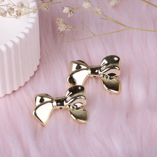 Pair of polished gold bow stud earrings on soft pink faux fur background with decorative dried flowers