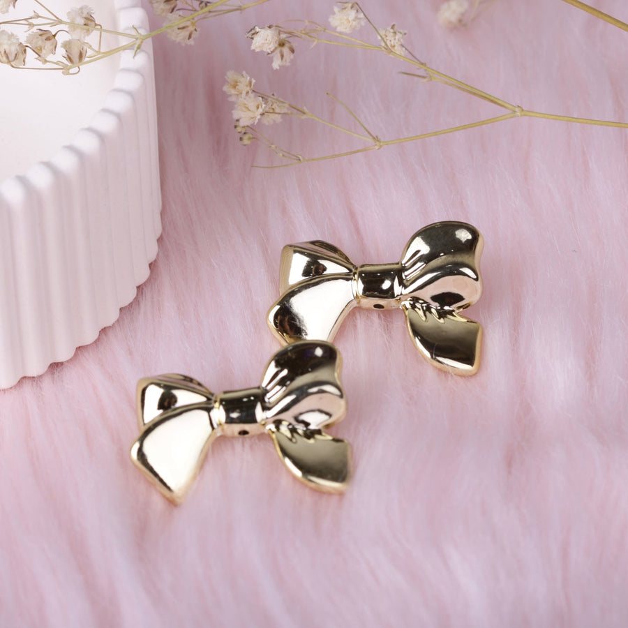 Pair of polished gold bow stud earrings on soft pink faux fur background with decorative dried flowers