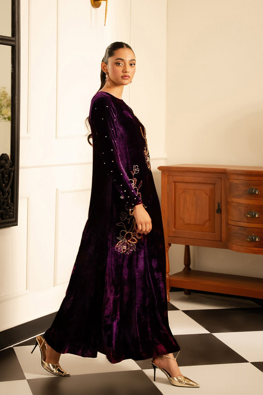 Side view of model in flowing deep purple velvet dress with crystal-embellished long sleeves and gold heels.