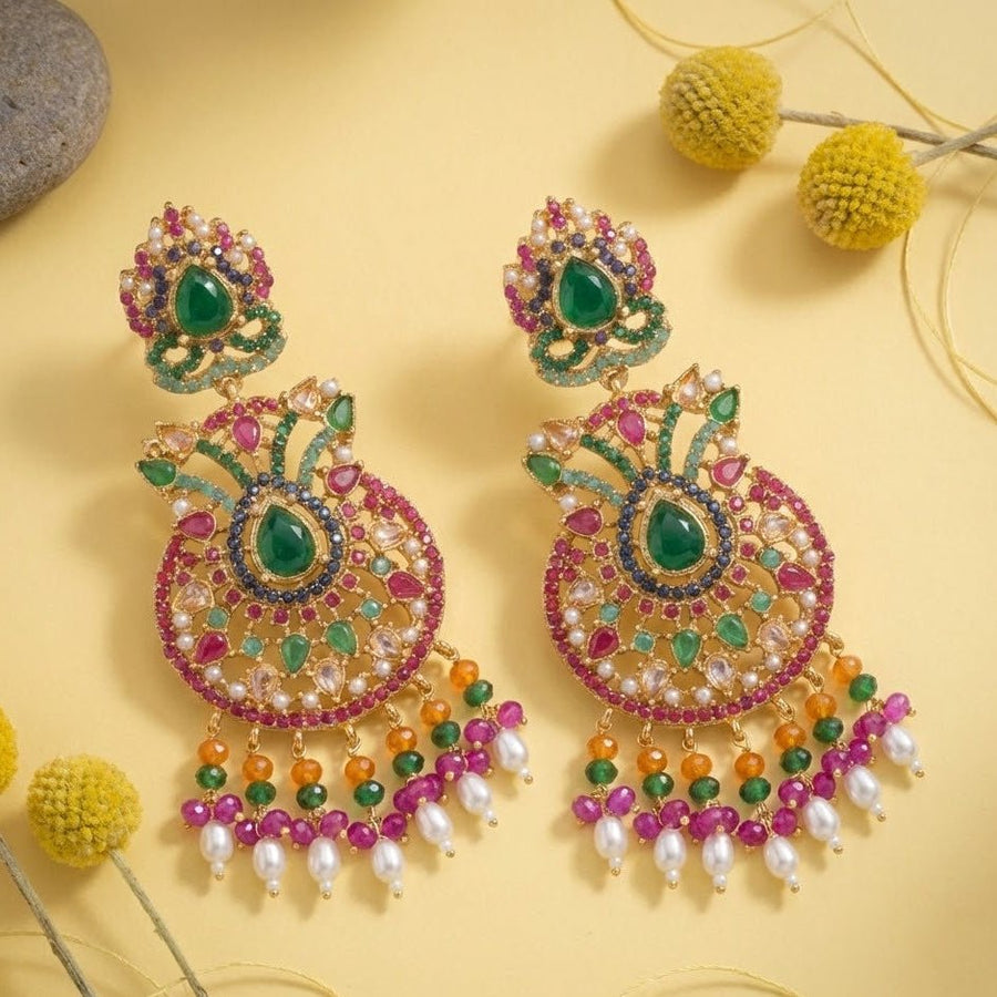 Colorful multi-gemstone jhumka earrings placed on pastel yellow surface with decorative yellow pom-pom flowers background