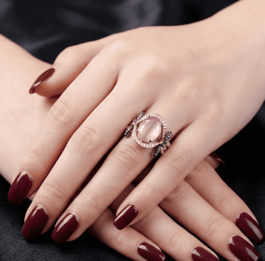 Elegant rose gold ring with oval pink semi-precious stone, clear halo stones, and black oxidized metal bows, modeled on hand