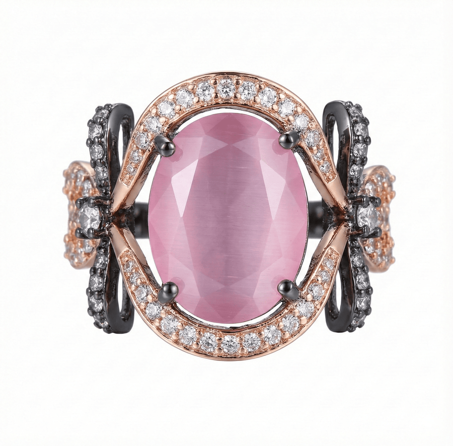 Close-up of a rose gold ring with a large oval pink semi-precious gemstone surrounded by small clear stones and black oxidized bow accents