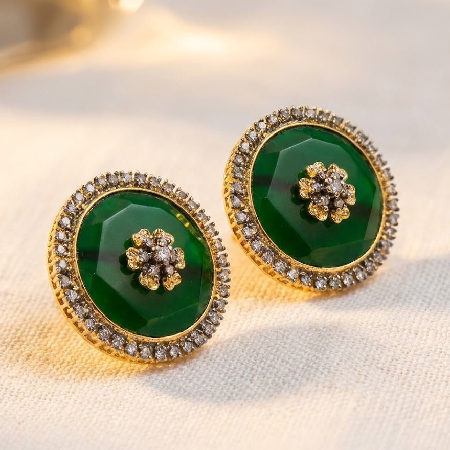Close-up of round stud earrings with emerald green faceted central stones surrounded by a halo of sparkling stones in gold-tone metal