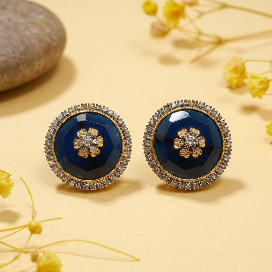 Pair of navy blue stud earrings with sparkling stone halos set on yellow surface with dried flowers as backdrop